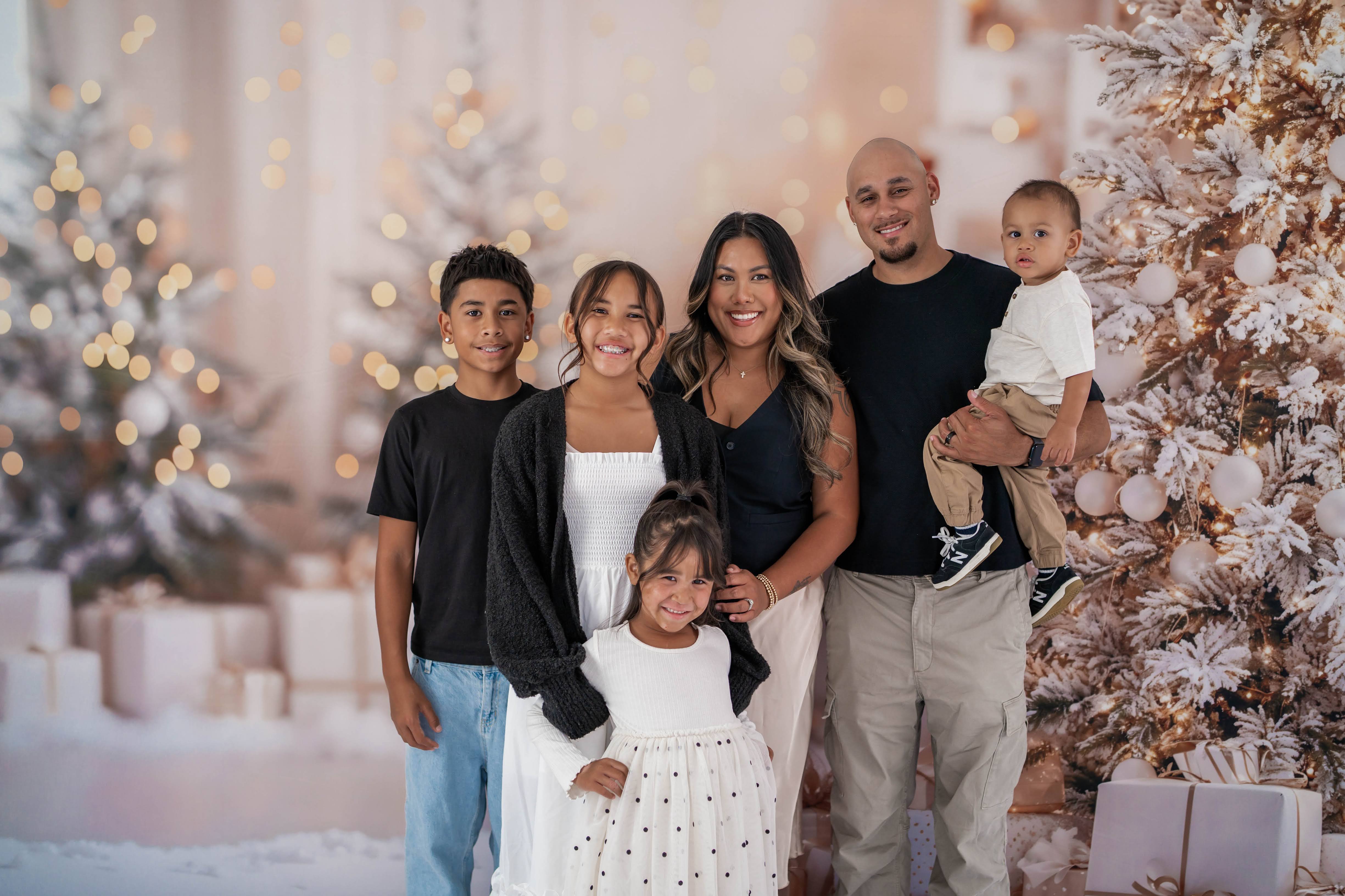 Family of six smiling together between flocked Christmas trees and stacked white gifts.