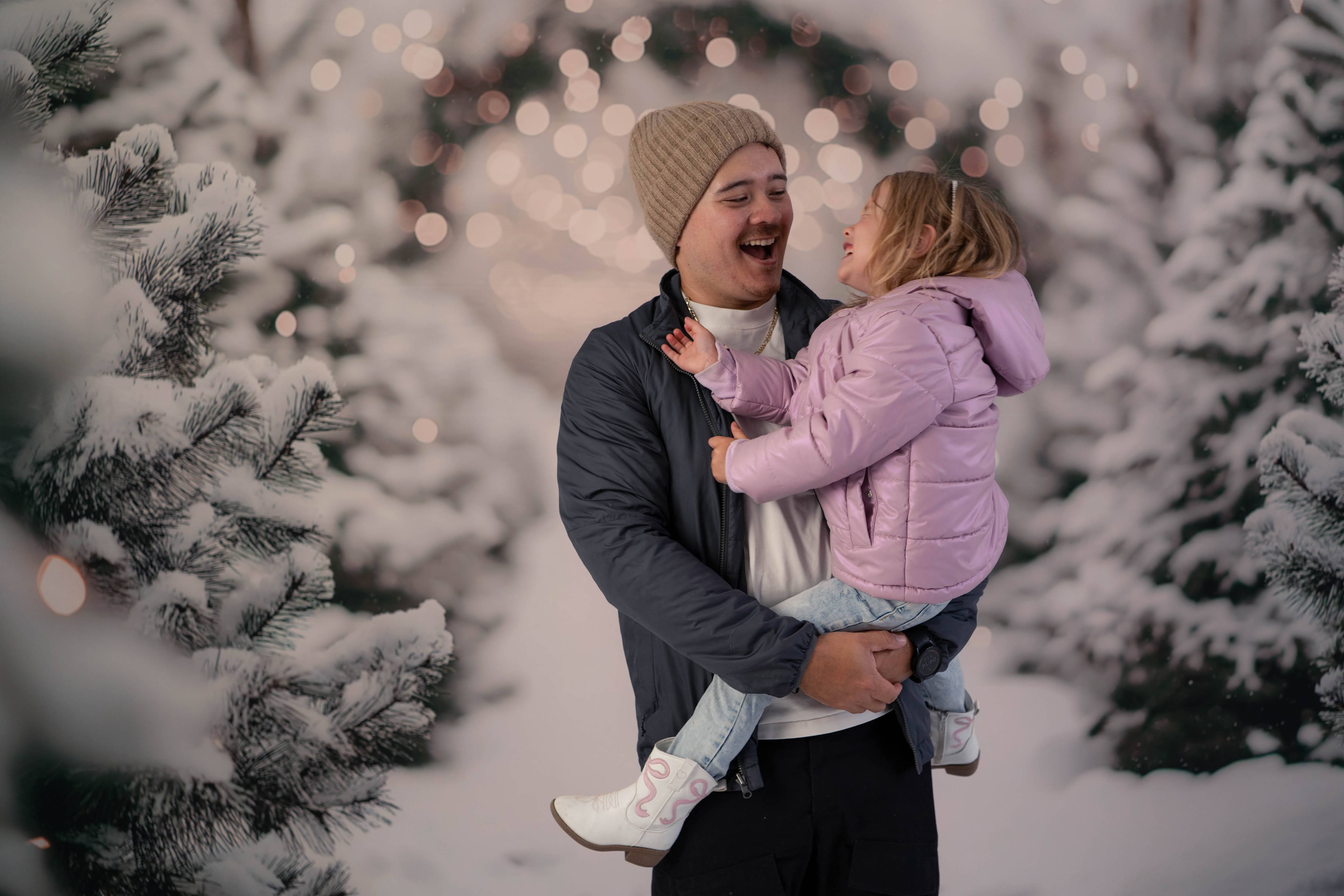Dad and his daughter laughing together among snowy evergreens and warm string lights.