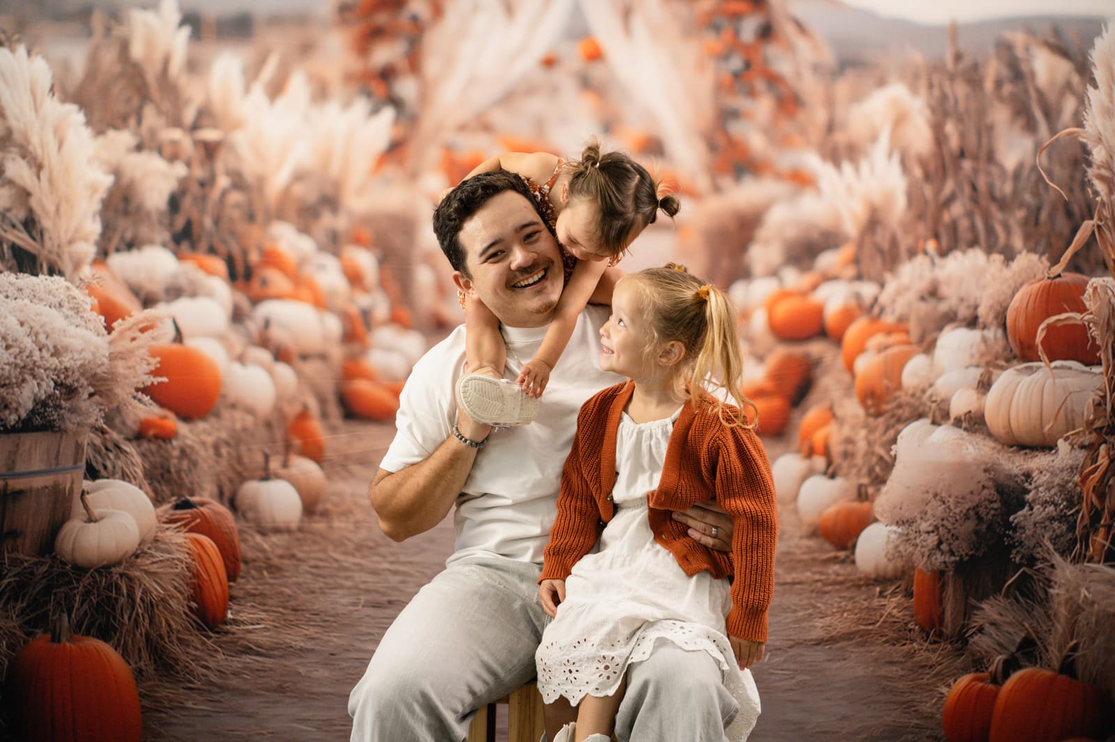Toddler leaning in to kiss her dad while her sister laughs in the pumpkin patch set.