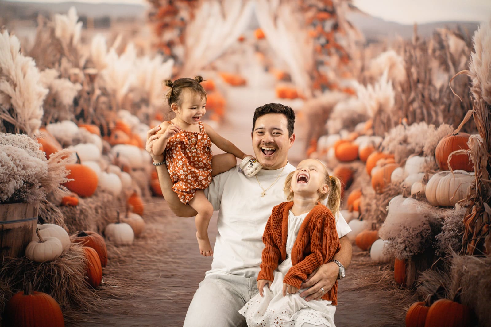 Father lifting his toddler as his older daughter laughs beside him among the pumpkins.