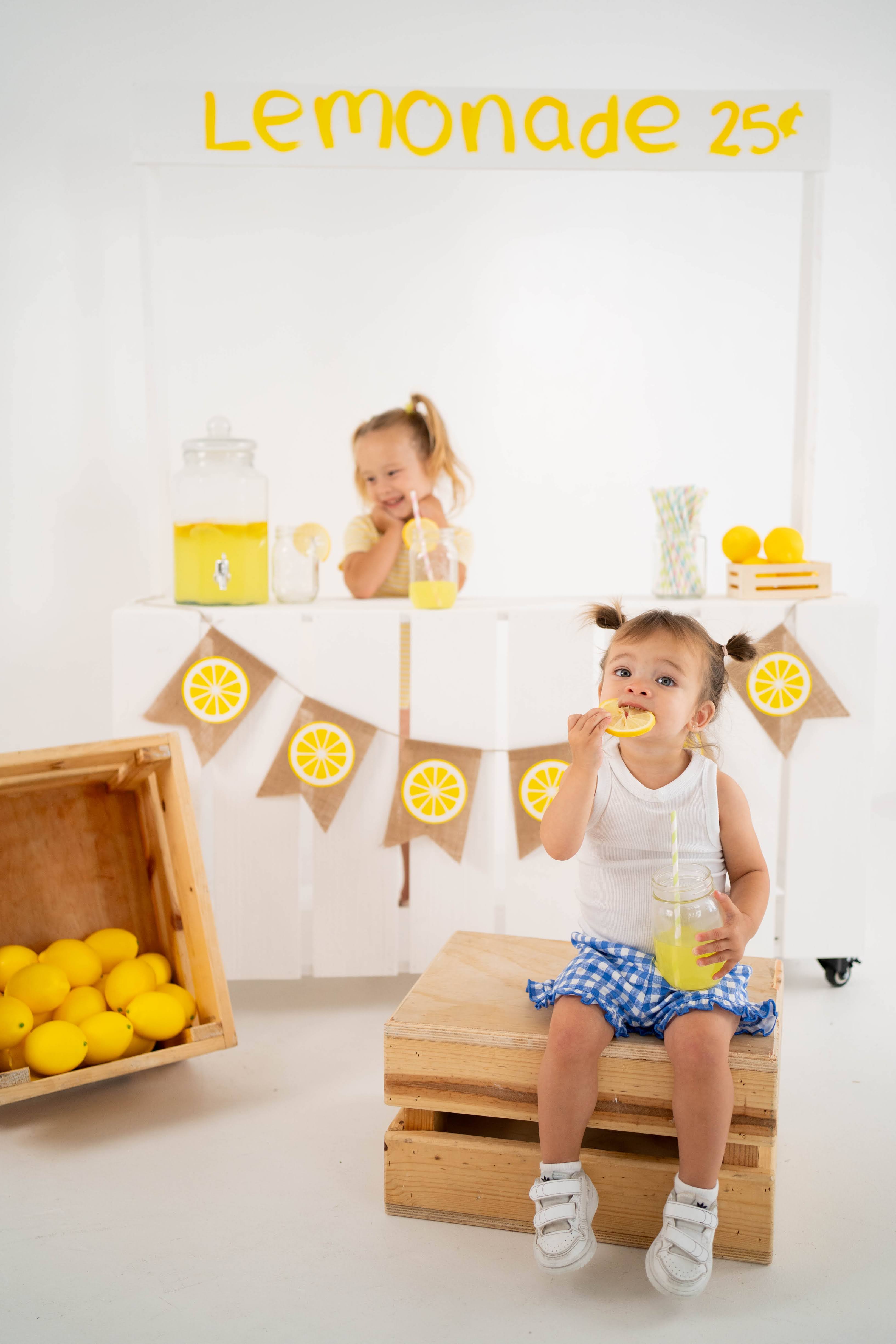 Toddler sipping lemonade on a wooden crate in the lemonade stand setup.