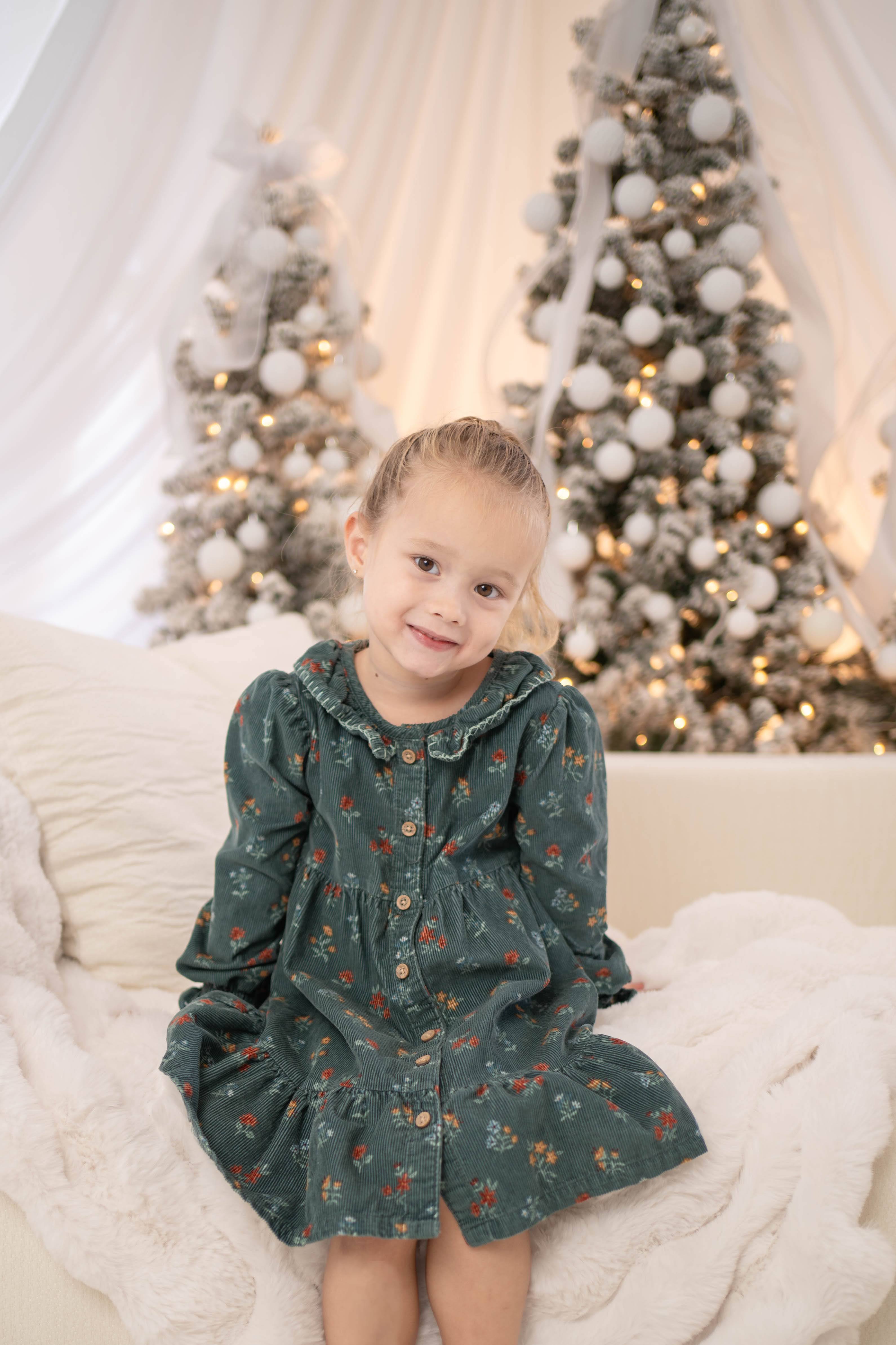 A young girl in a dark green floral dress sits smiling on a plush white chair in front of a softly lit Christmas tree decorated with white baubles.