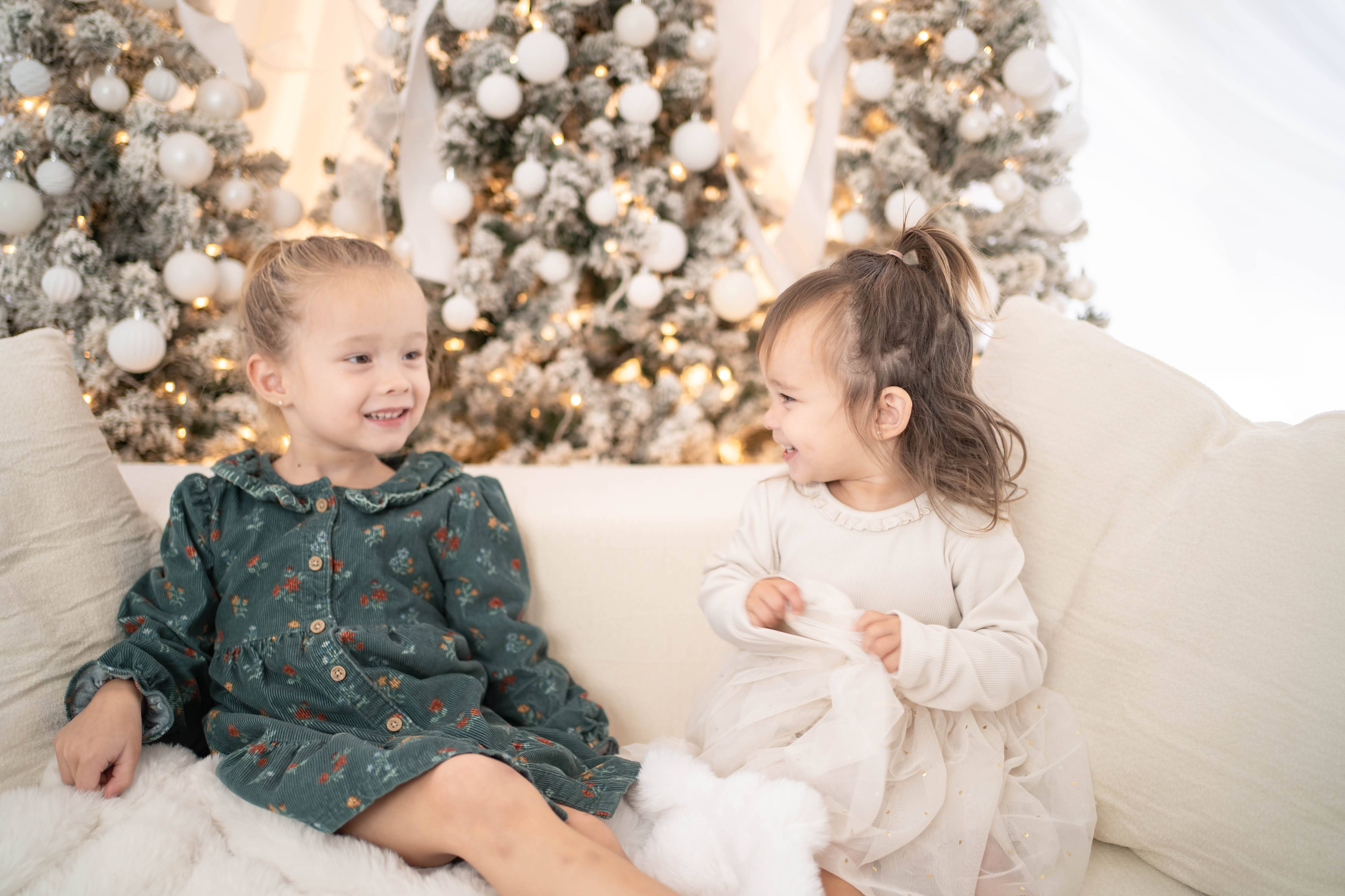 Two young children sit close together on a pale sofa in front of a snowy, warmly lit Christmas tree, smiling at each other.
